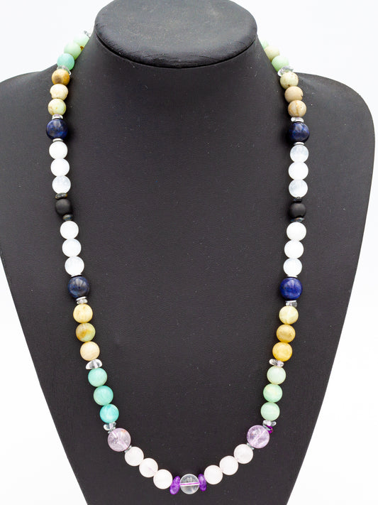 A handcrafted beaded necklace featuring an array of vibrant gemstones, each resonating with the energy of a specific element and chakra. The beads transition seamlessly from deep grounding hues to ethereal whites and blues, symbolizing the interconnectedness of earth, fire, water, air, and spirit. Set against a sleek black display, the necklace radiates an aura of balance, alignment, and spiritual attunement