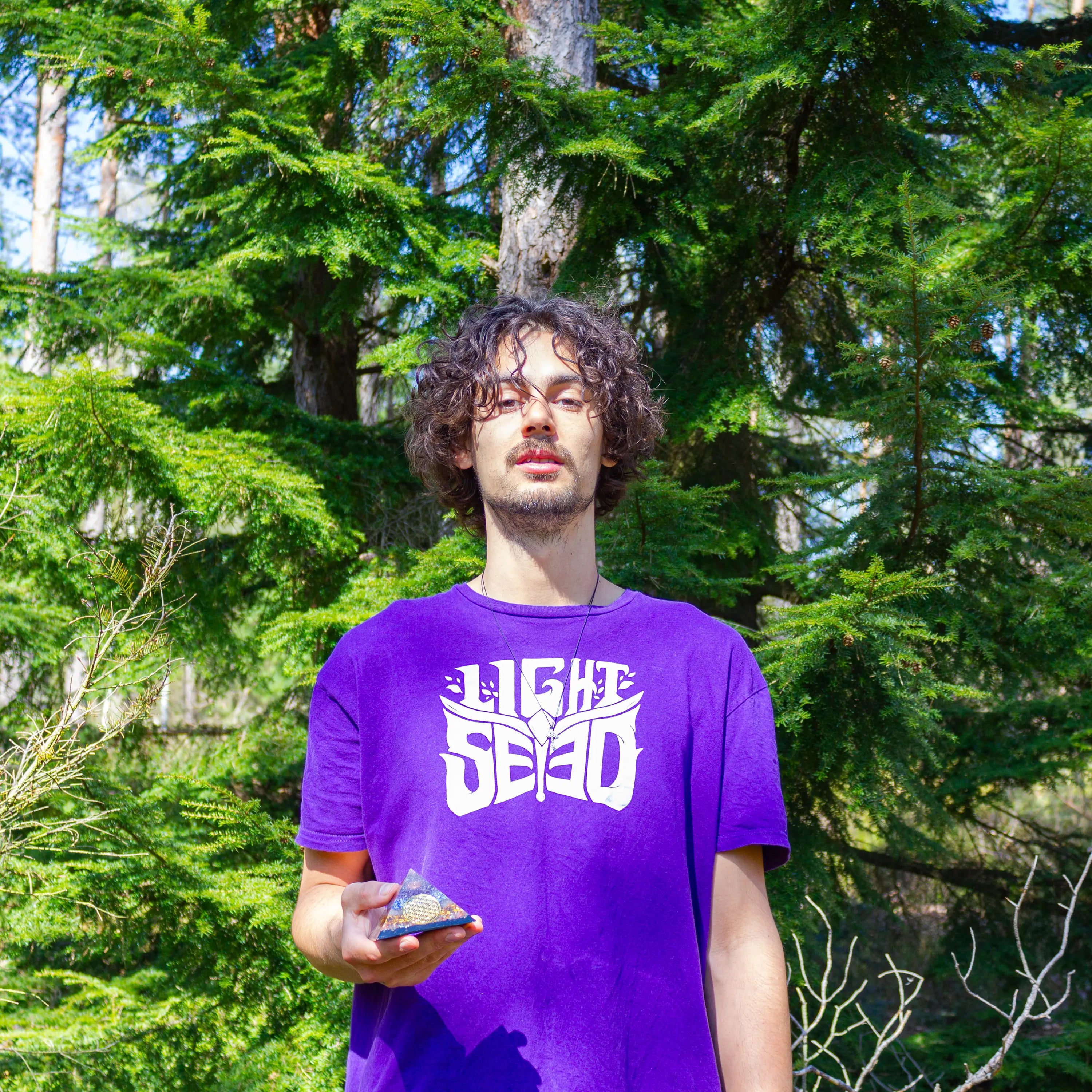 A radiant individual wearing a purple "Lightseed" shirt stands in front of vibrant pine trees, holding a glowing orgonite pyramid in his left hand. The scene exudes natural energy and clarity, with the pyramid amplifying the high-frequency transmission from forest to soul. A perfect embodiment of Lightseed’s mission to ground cosmic energy through sacred tools and nature's presence.